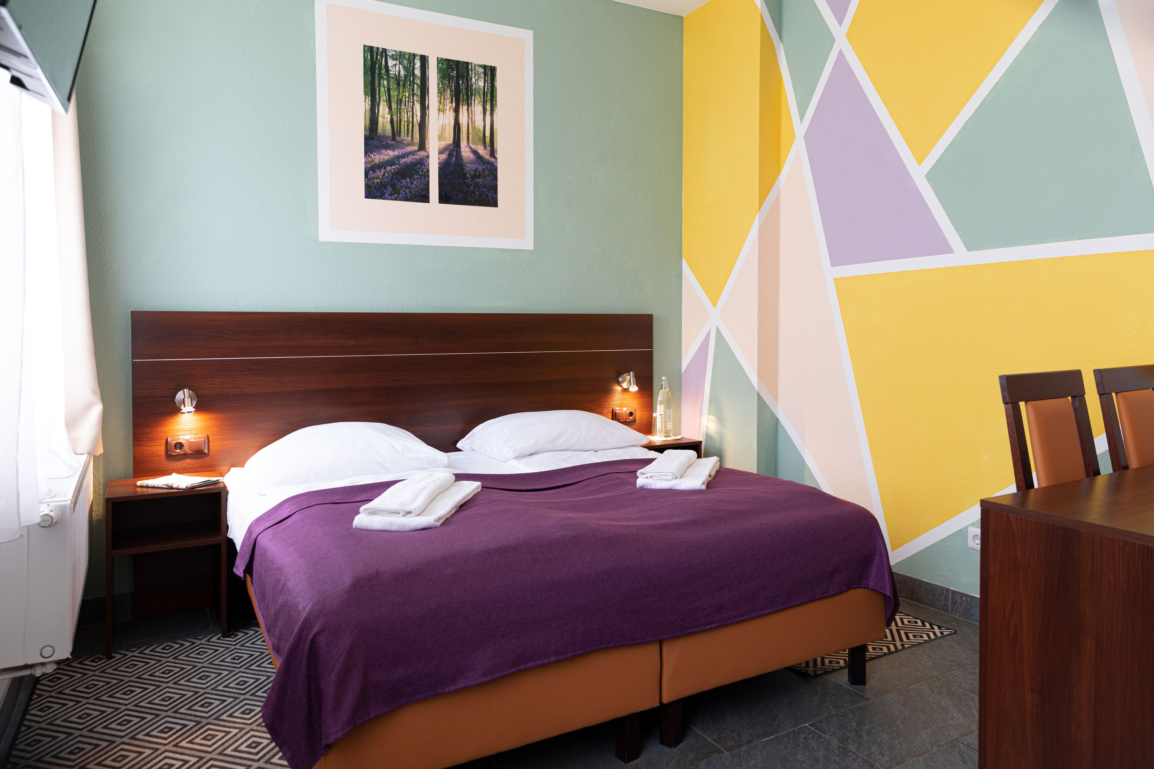 Ein ordentlich bezogenes Bett mit einer violetten Decke, zwei weißen Kissen und einem hölzernen Kopfteil. Es gibt zwei Nachttische mit Lampen, ein gerahmtes Bild an der Wand und rechts ein kleiner Tisch mit Stühlen.