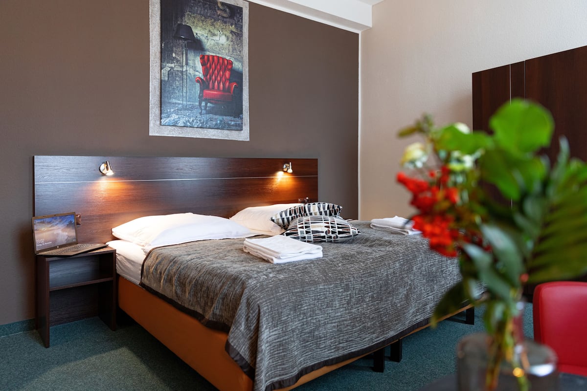 Una cama bien hecha con una manta gris, dos lámparas de noche, una mesita con un ordenador portátil y una silla roja en la esquina.