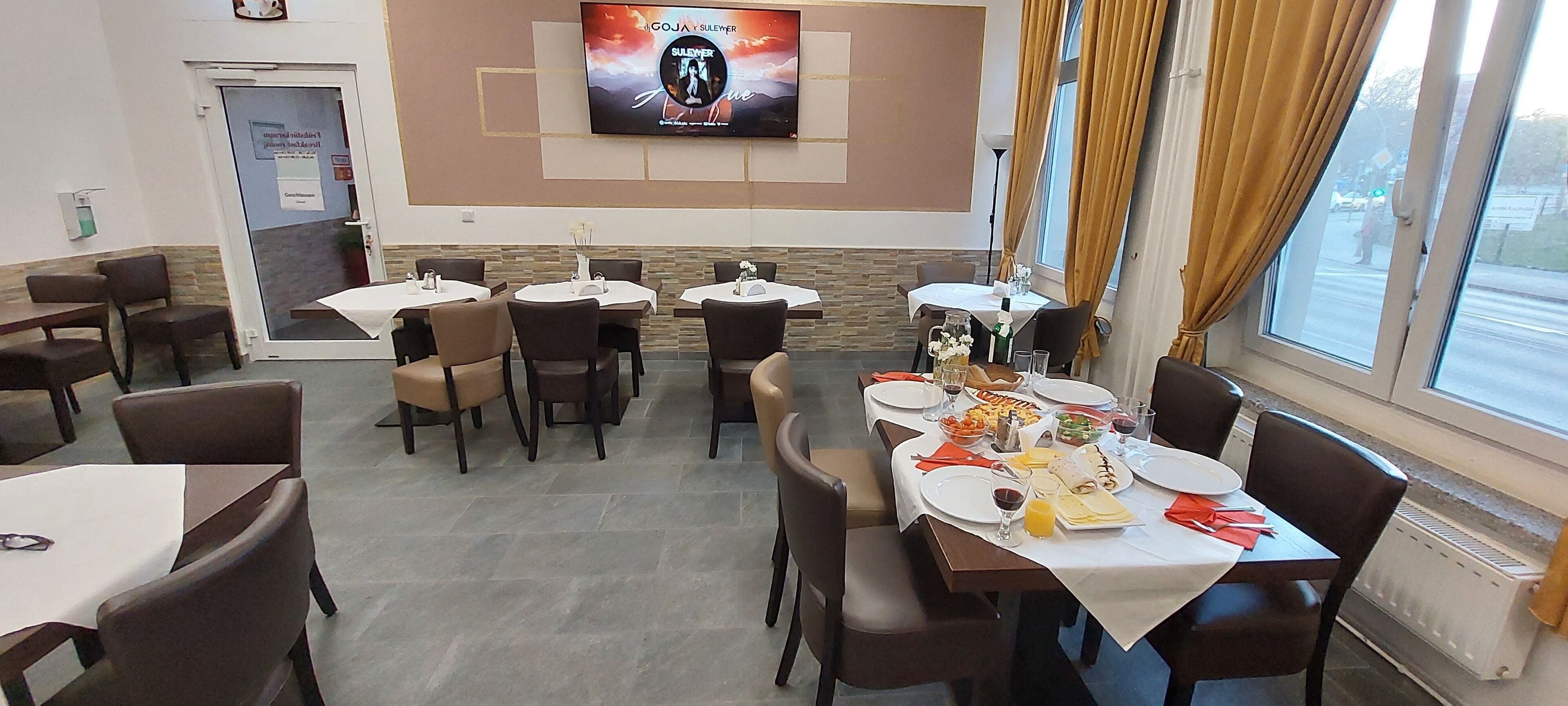 Ein Restaurant mit mehreren Tischen und Stühlen, einem an der Wand befestigten Fernseher und einem Fenster mit Vorhängen.