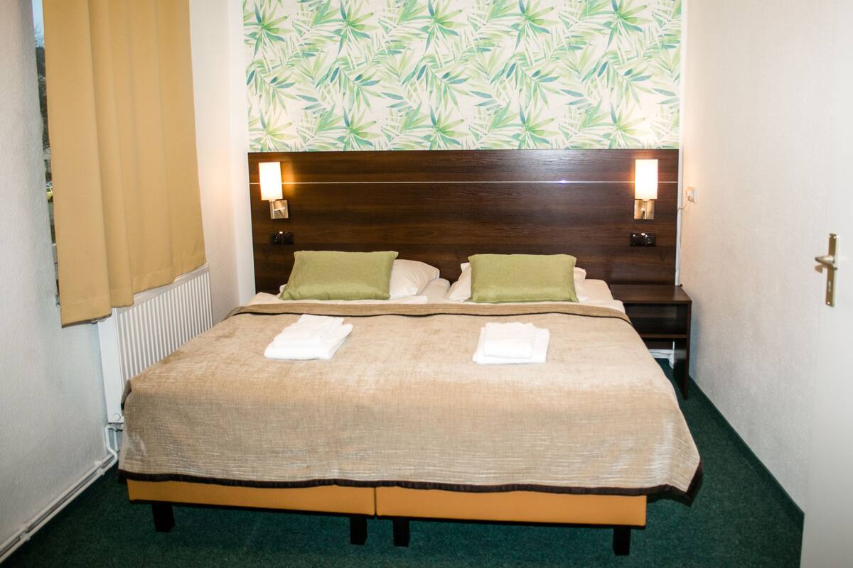 Una cama bien tendida con un cabecero marrón, dos lámparas de noche y un papel tapiz con estampado de hojas tropicales.
