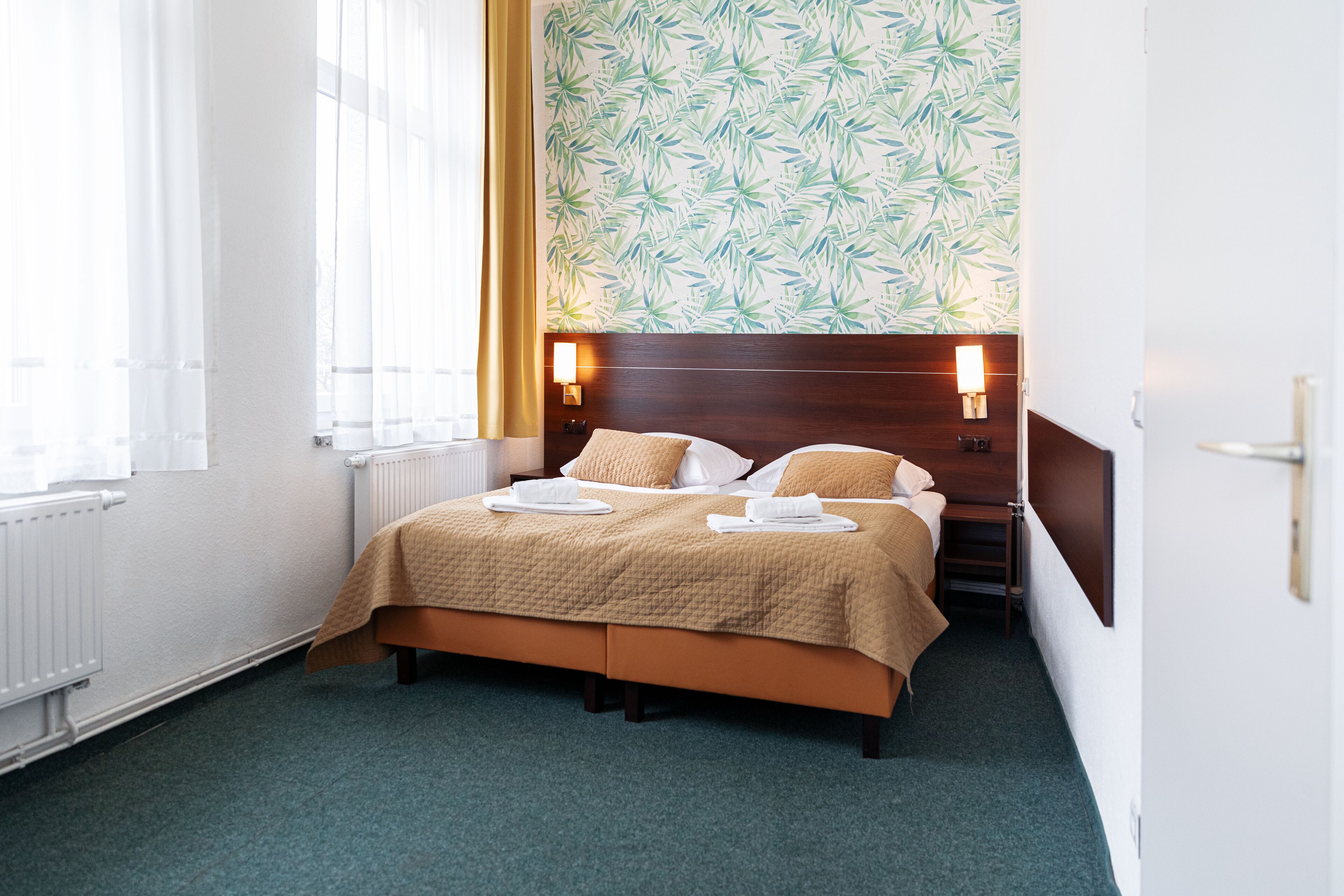Ein Hotelzimmer mit Bett, Nachttischlampen, einem Fenster mit Vorhängen und einer Wand mit Blättermuster.