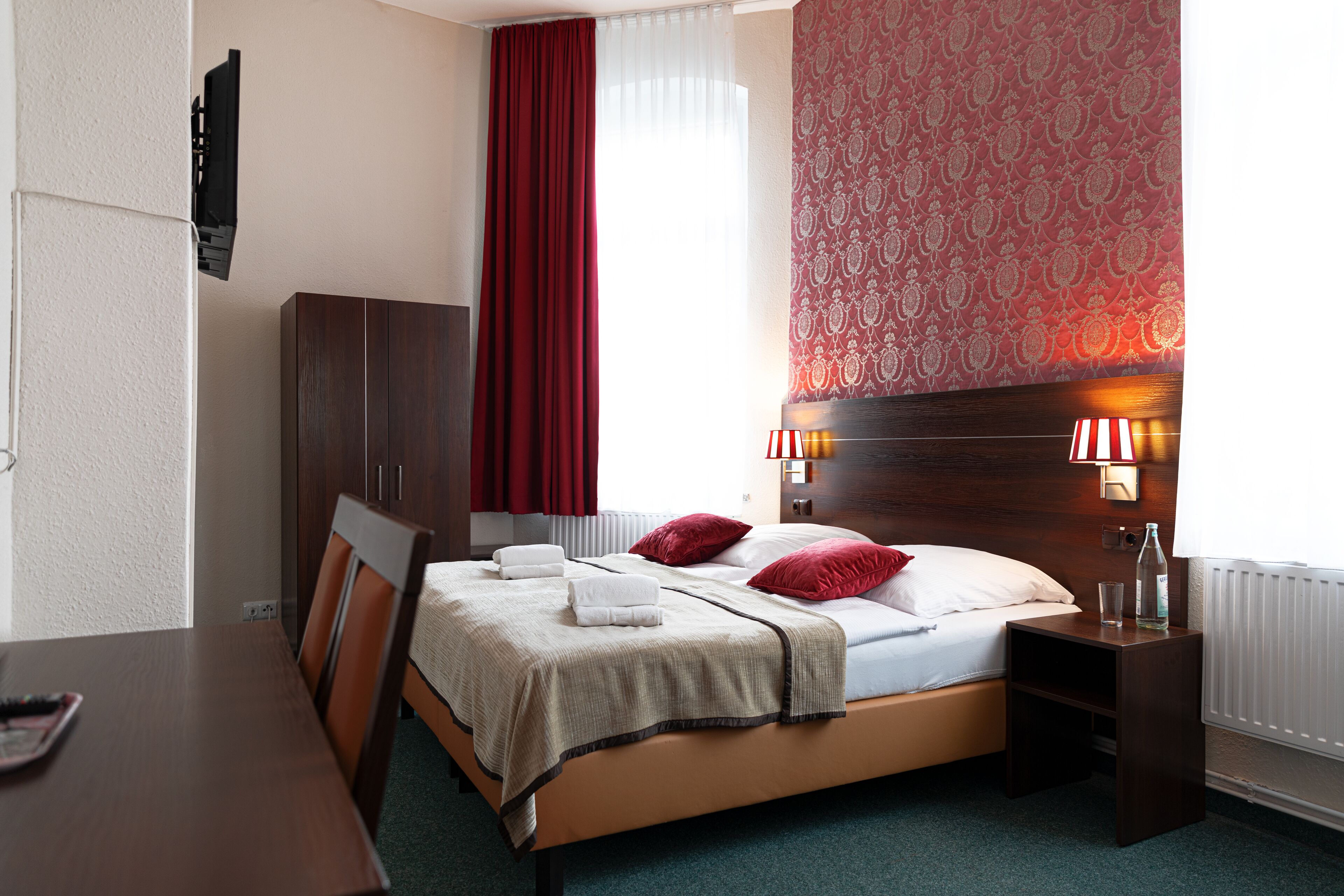 Une chambre d’hôtel comprenant un lit, un bureau, une chaise, une armoire, une table de chevet avec une lampe et une fenêtre avec des rideaux.