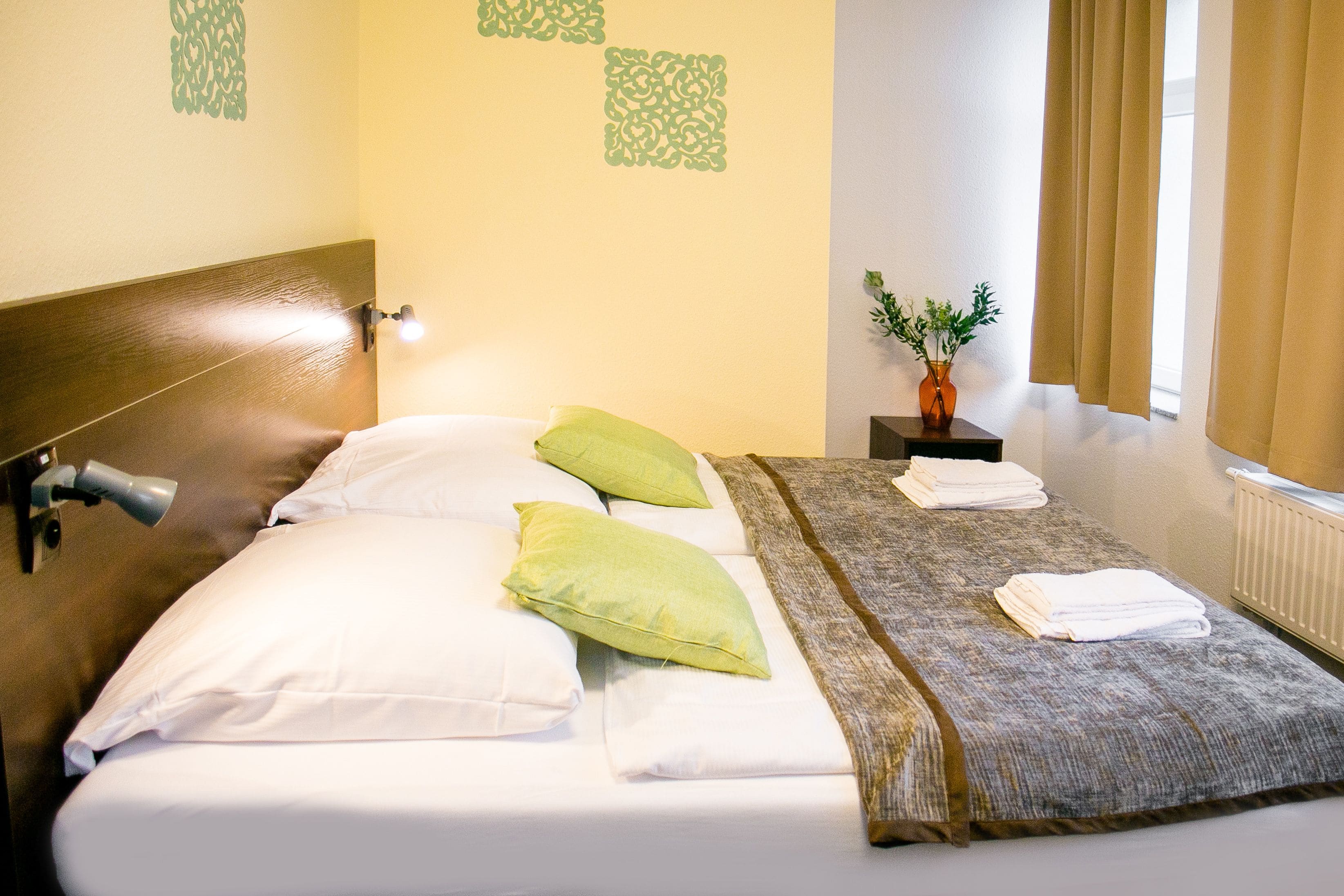 A neatly made bed with white and green pillows, a gray blanket, and a small table with a vase of flowers.