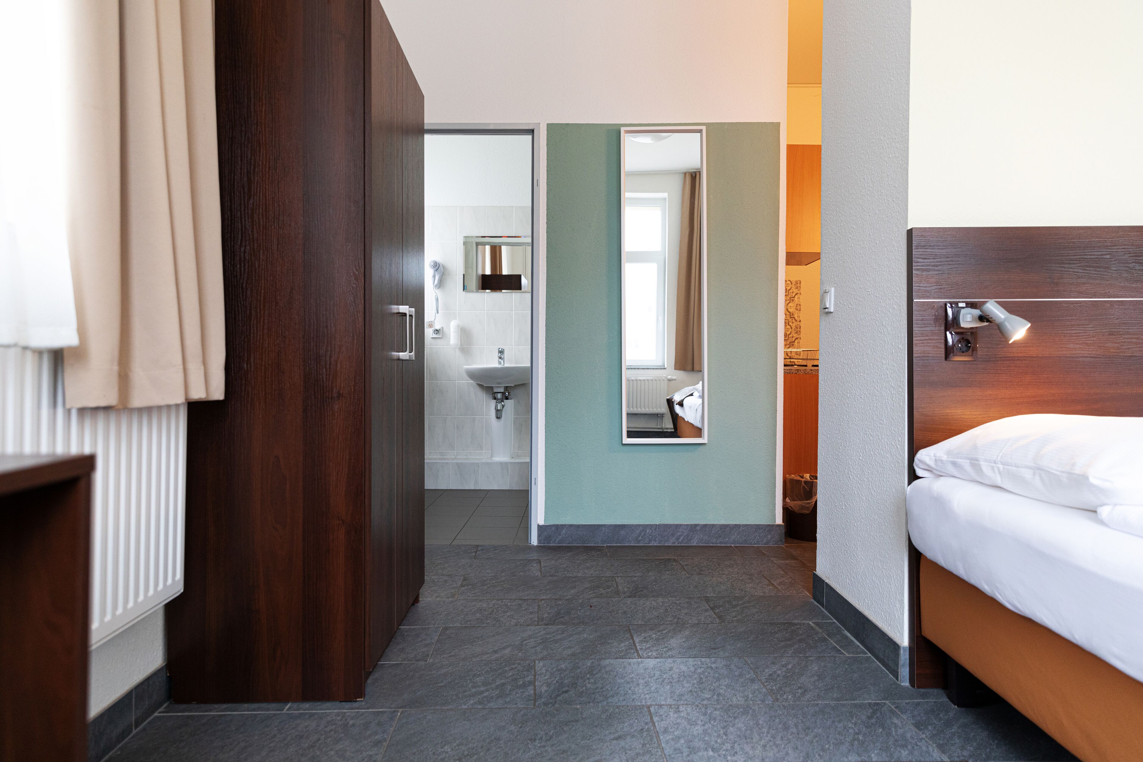 Habitación de hotel con armario de madera, cama con ropa blanca, baño con lavamanos y espejo, y suelo de baldosas.