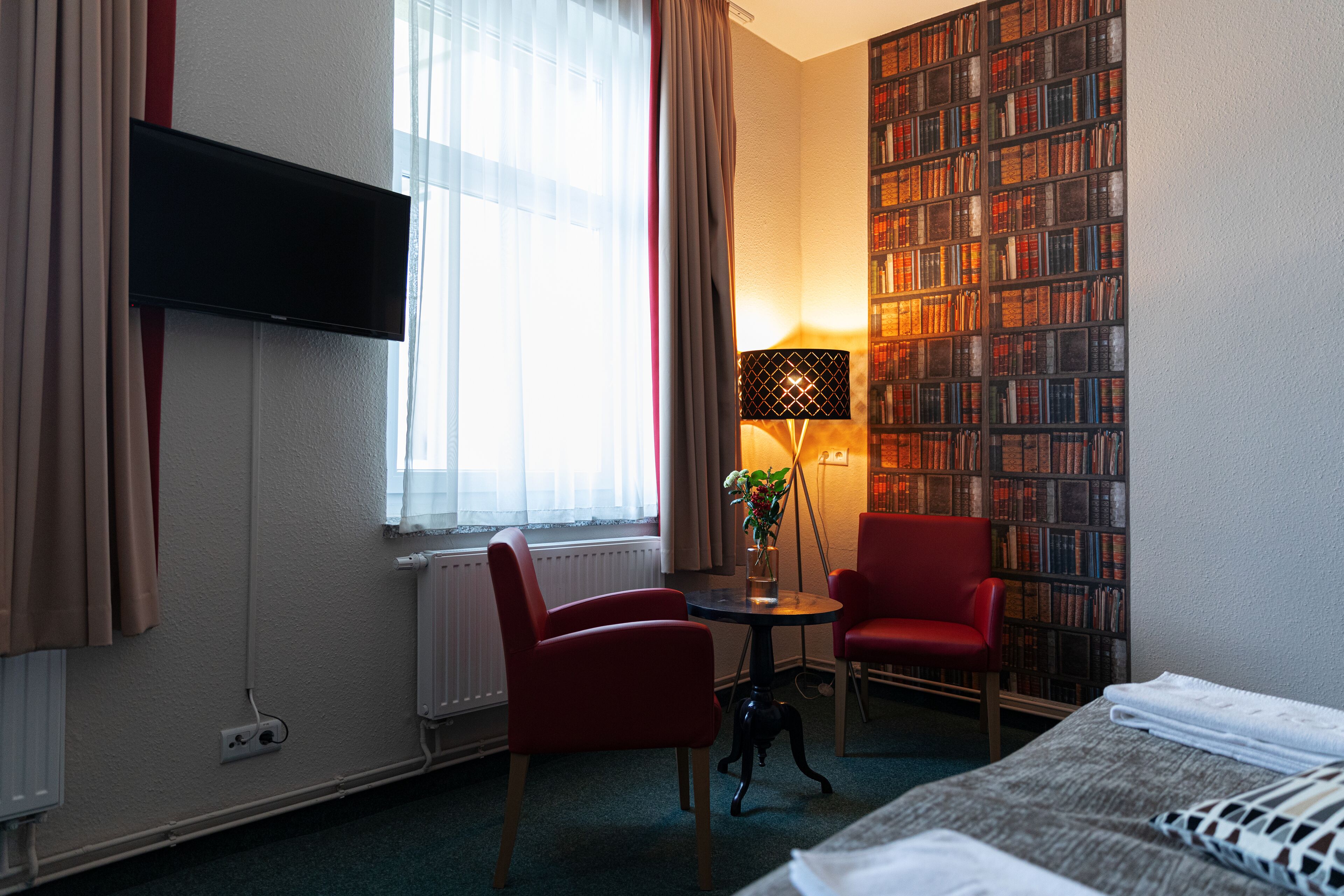 Une chambre d’hôtel avec un lit, un fauteuil rouge, une petite table avec une lampe, une bibliothèque et un téléviseur fixé au mur.
