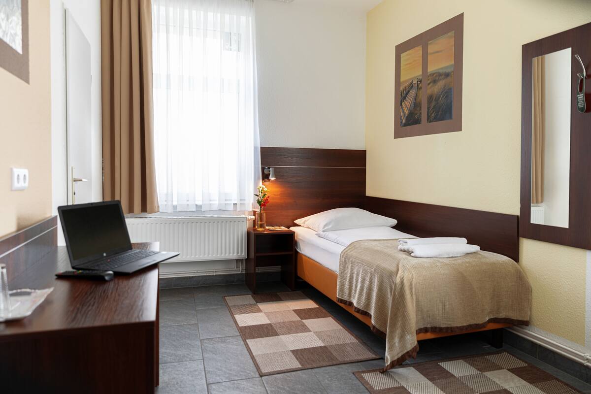 Una habitación de hotel con cama, escritorio con laptop, mesita de noche, ventana con cortinas y un cuadro en la pared.
