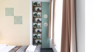 Een hotelkamer met een bed, een houten plank met planten en boeken, een raam met gordijnen en een muur met decoratieve patronen.