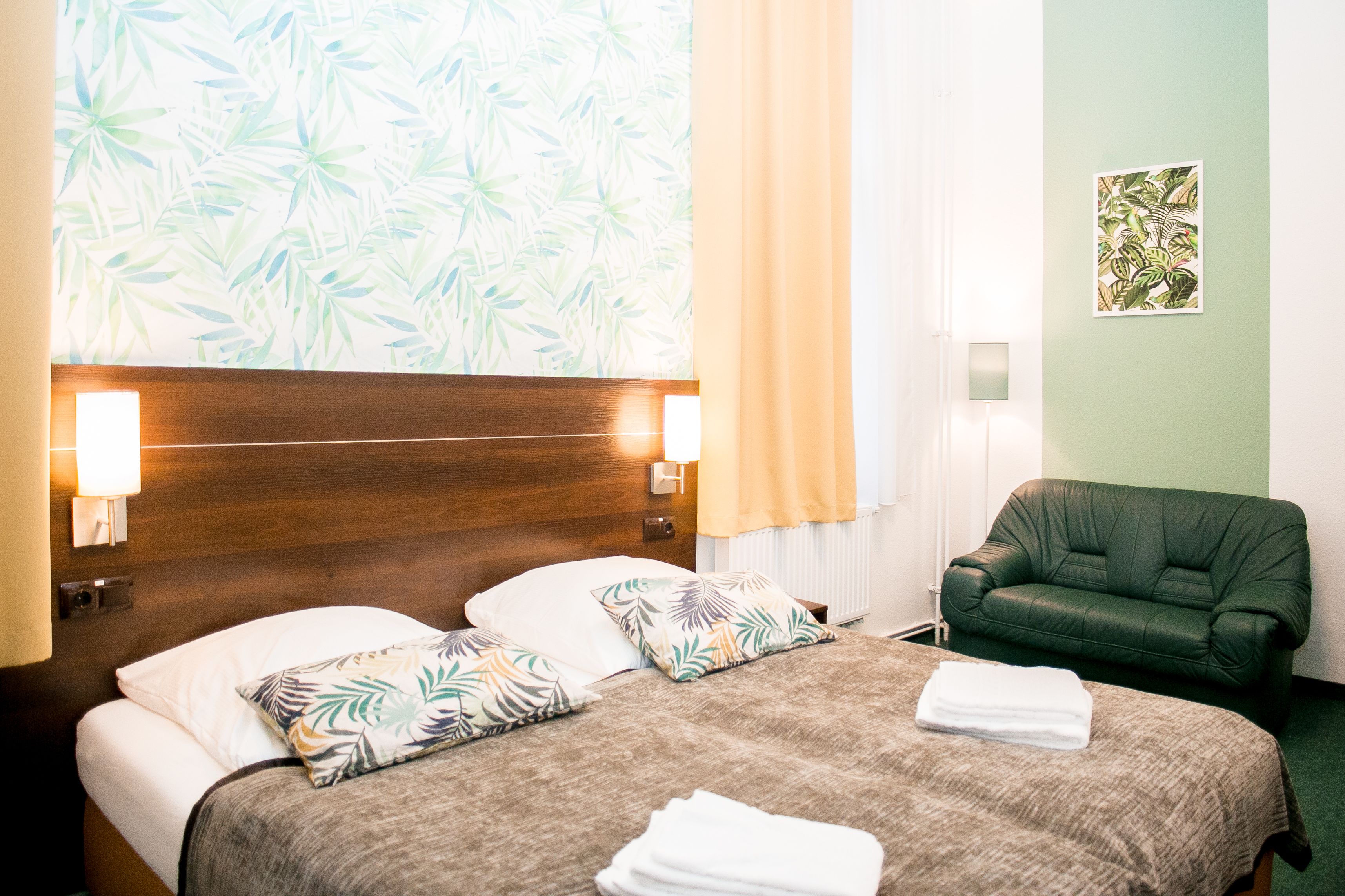 A hotel room with two beds, a green armchair, a tropical wallpaper, and a framed plant artwork on the wall.