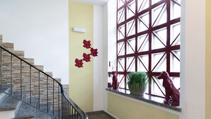 Una escalera con escalones de piedra y barandilla negra, una pared con diseño de ventana geométrico y una escultura roja en el alféizar.