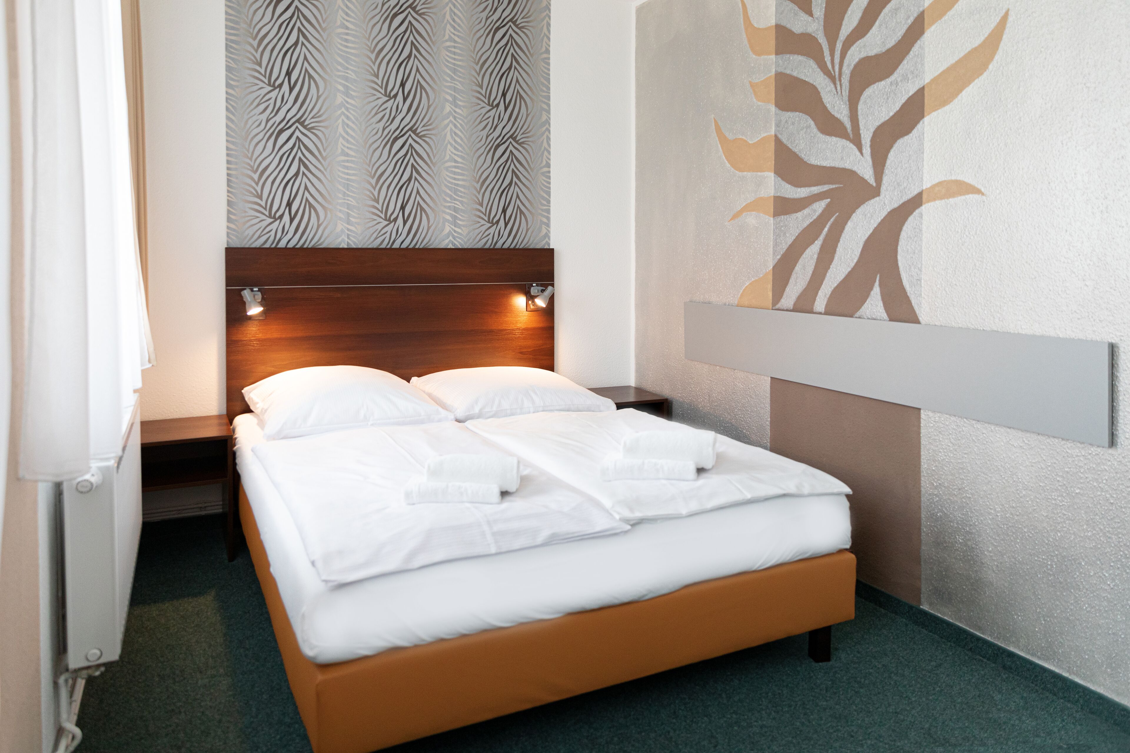 Una cama bien tendida con sábanas blancas, cabecera de madera y pared con un diseño de hojas.