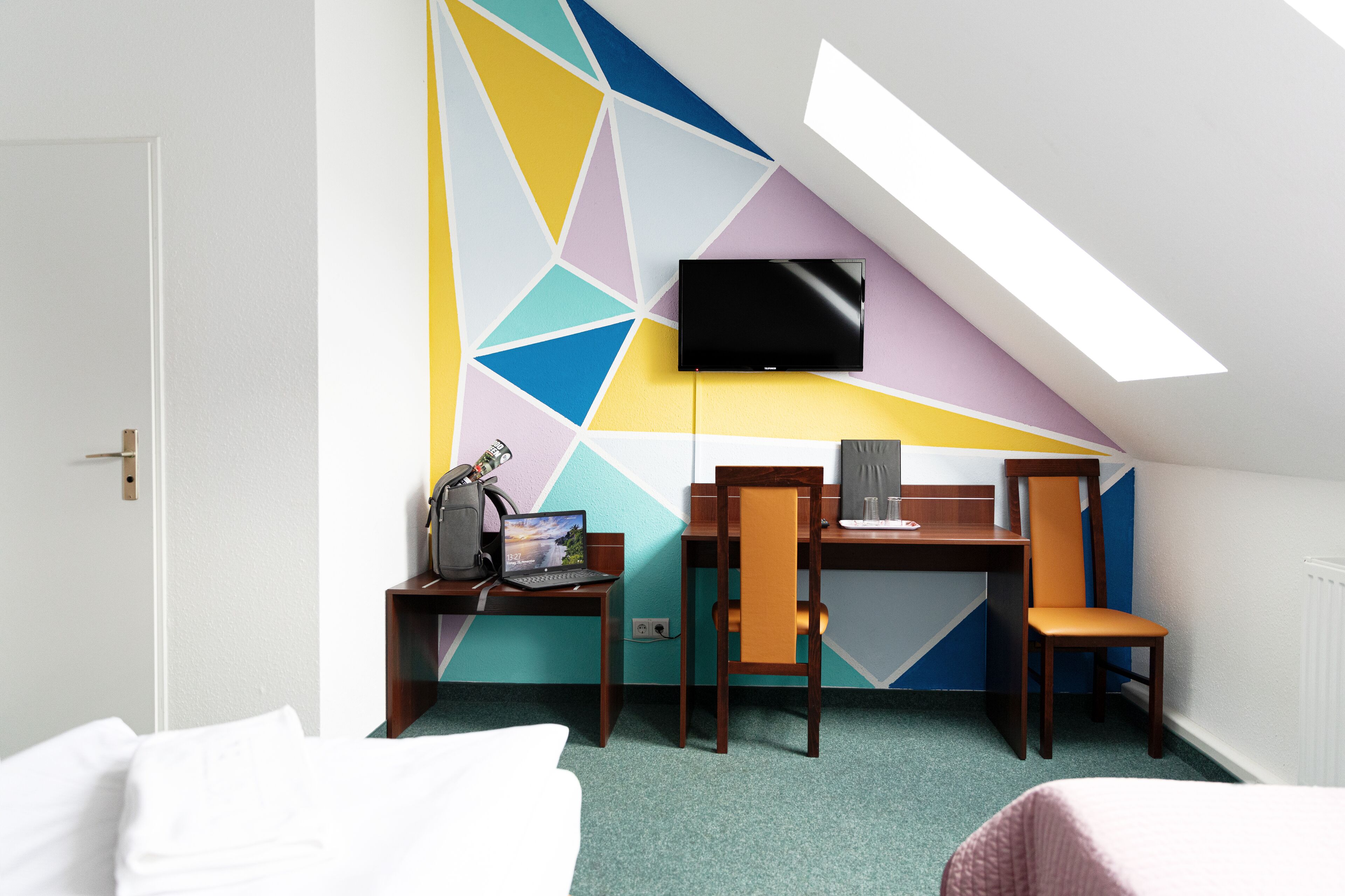 A hotel room with a sloped ceiling, a flat-screen TV mounted on the wall, a desk with a laptop, two chairs, and a bed with white bedding.