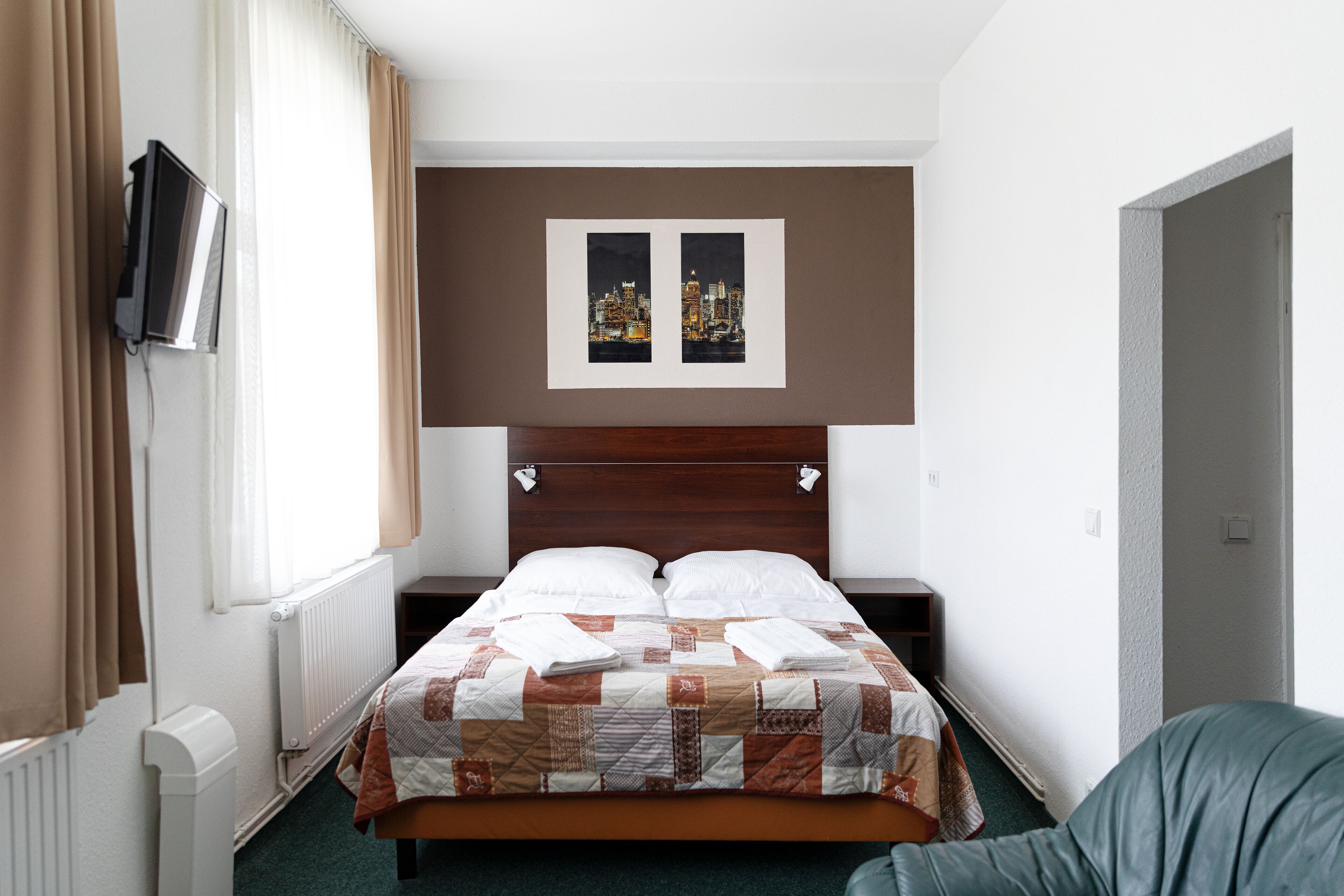 Habitación de hotel con cama, televisor montado en la pared, ventana con cortinas y una imagen de un paisaje urbano sobre la cama.