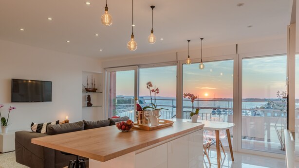 A modern kitchen with a wooden island, large windows with a view of the sunset, and a flat-screen TV mounted on the wall. - Amazing View Apartment (Zadar)