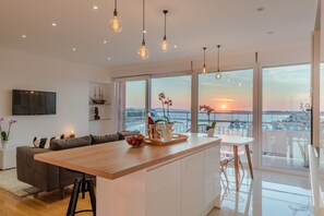 Una cocina moderna con una isla de madera, ventanas amplias con vistas al atardecer y un televisor de pantalla plana montado en la pared.