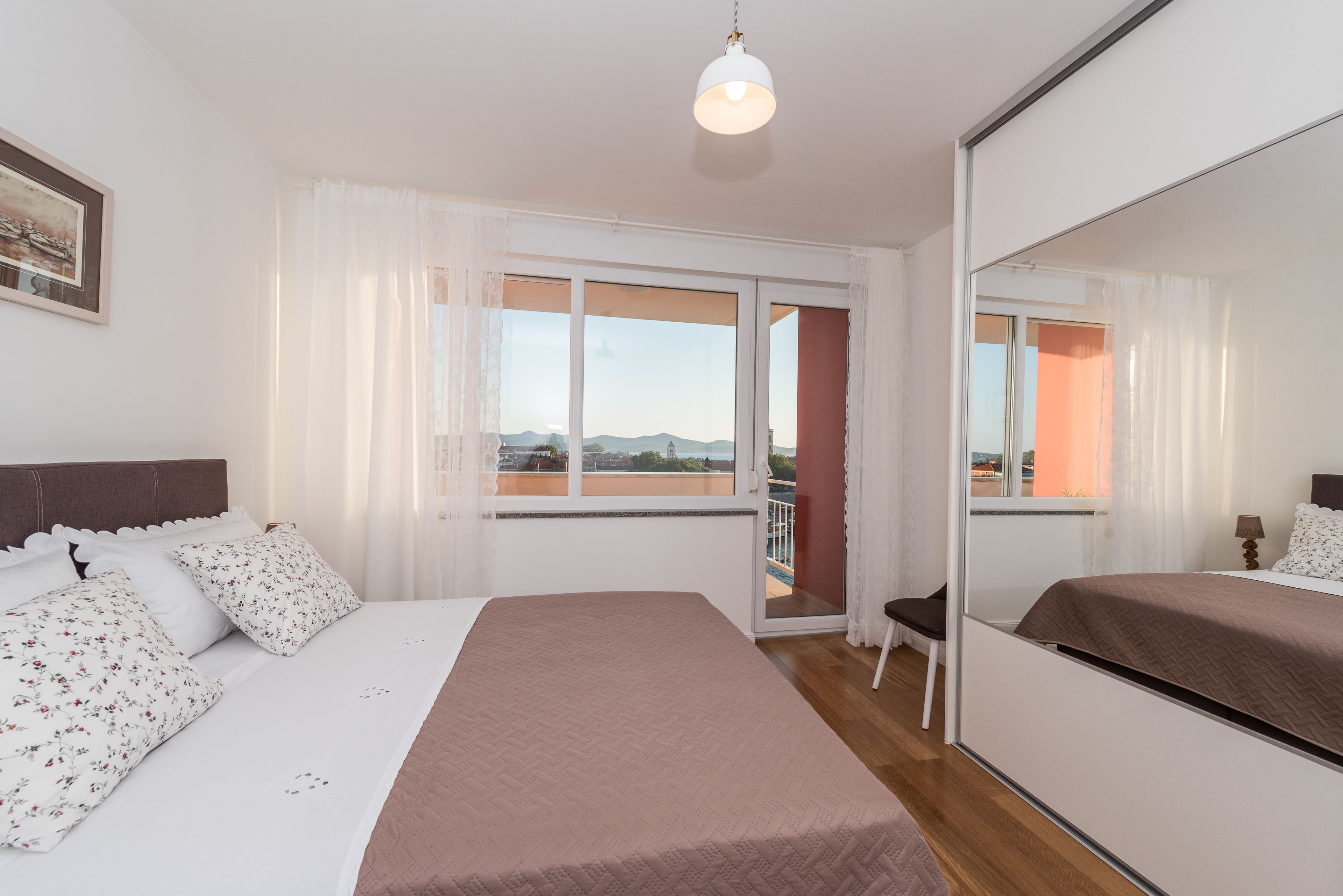 Un dormitorio con cama, ventana con cortinas, balcón y espejo.