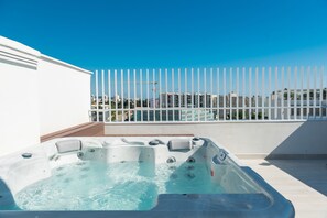A hot tub on a rooftop terrace with a white fence and buildings in the background.