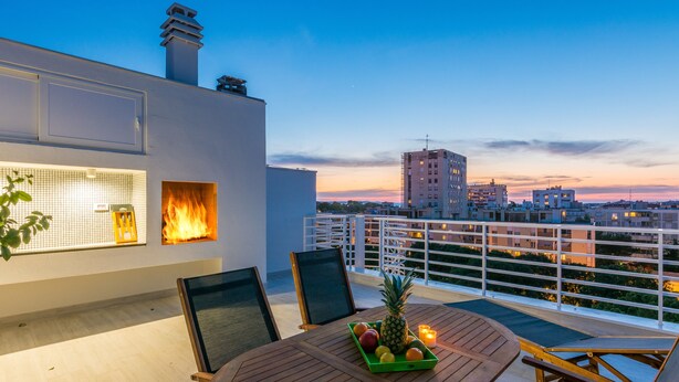 Une terrasse sur le toit, avec une table, des chaises, un foyer et une vue sur le paysage urbain au crépuscule.