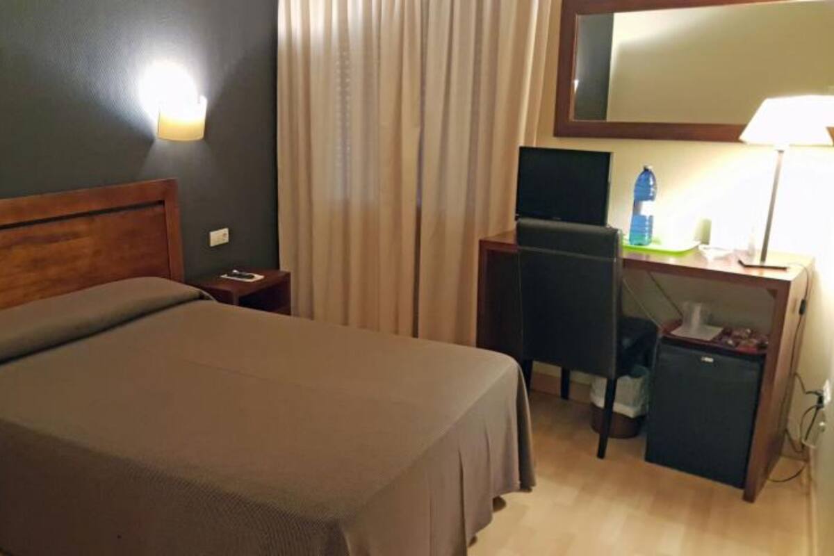 A hotel room with a bed, a desk, a chair, a lamp, and a mirror.