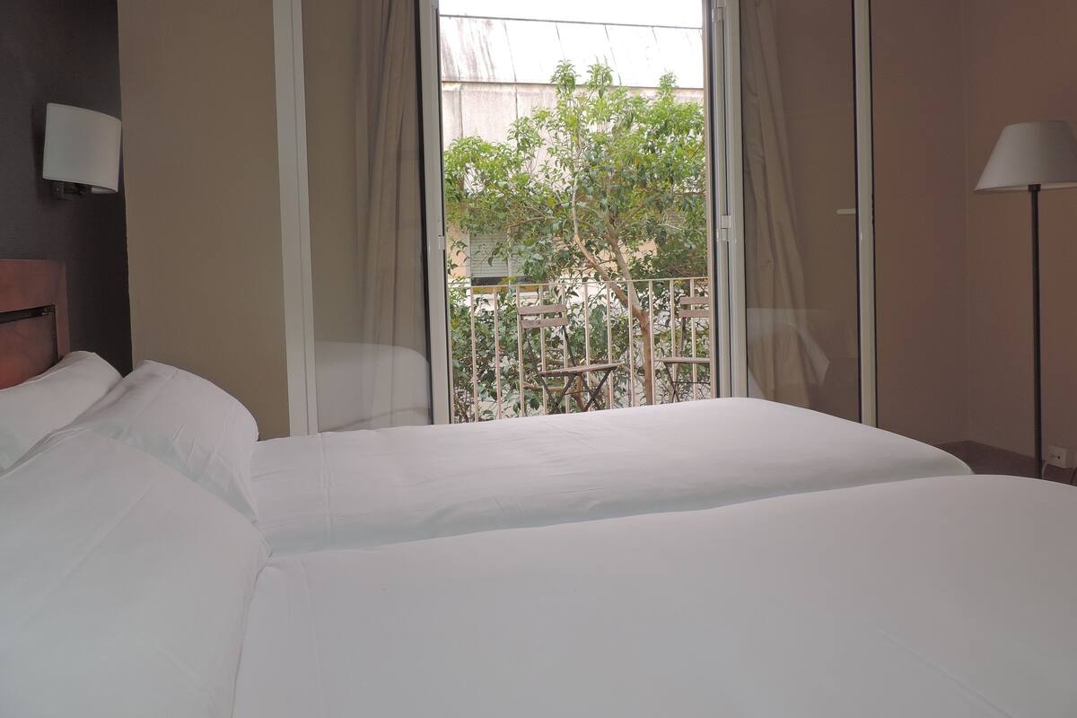 A hotel room with a large bed, a nightstand, a lamp, and a view of a balcony with trees and a fence.