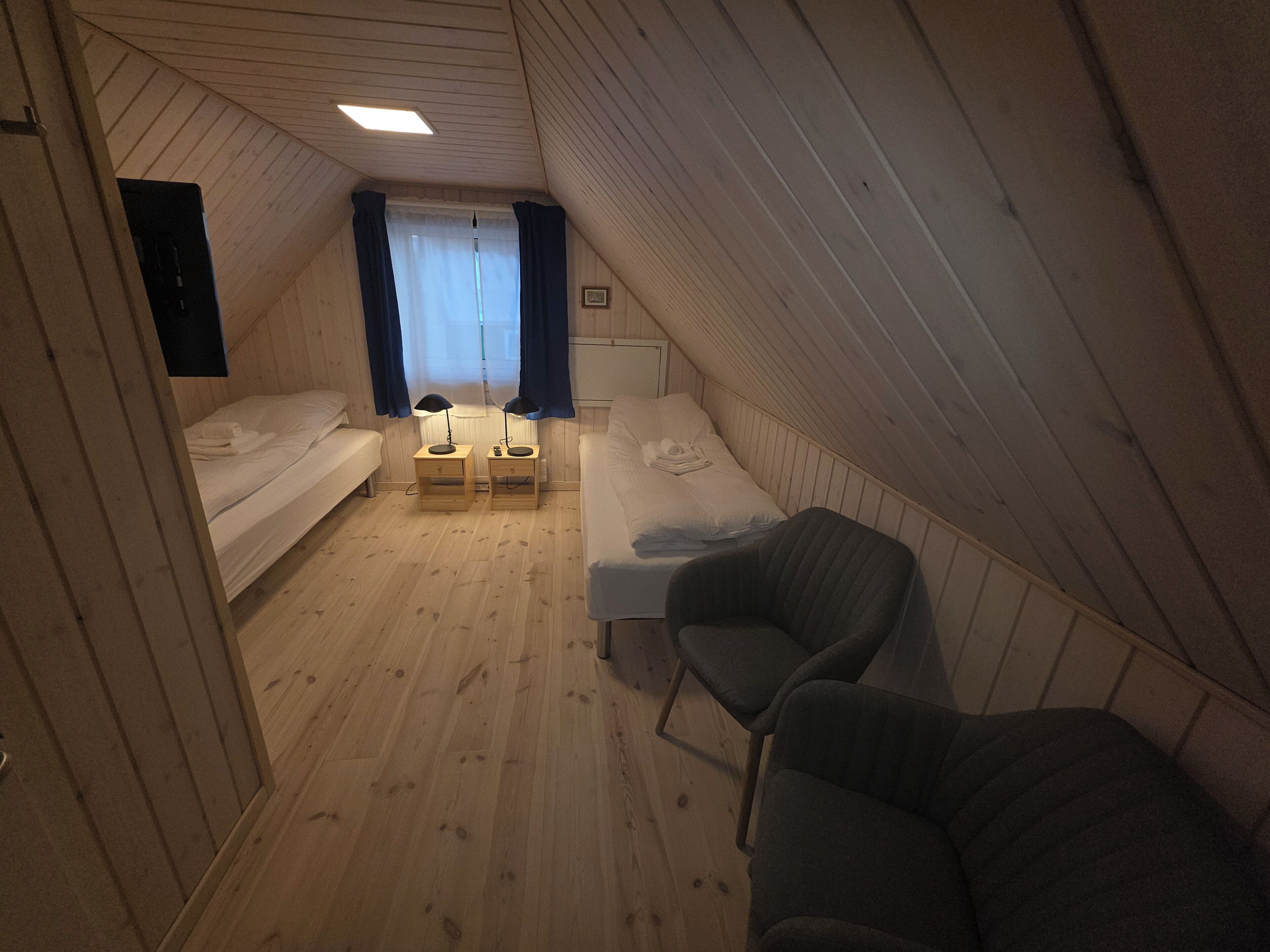 Une petite pièce aux murs lambrissés, comprenant deux lits simples, un bureau avec deux chaises et une fenêtre avec des rideaux.