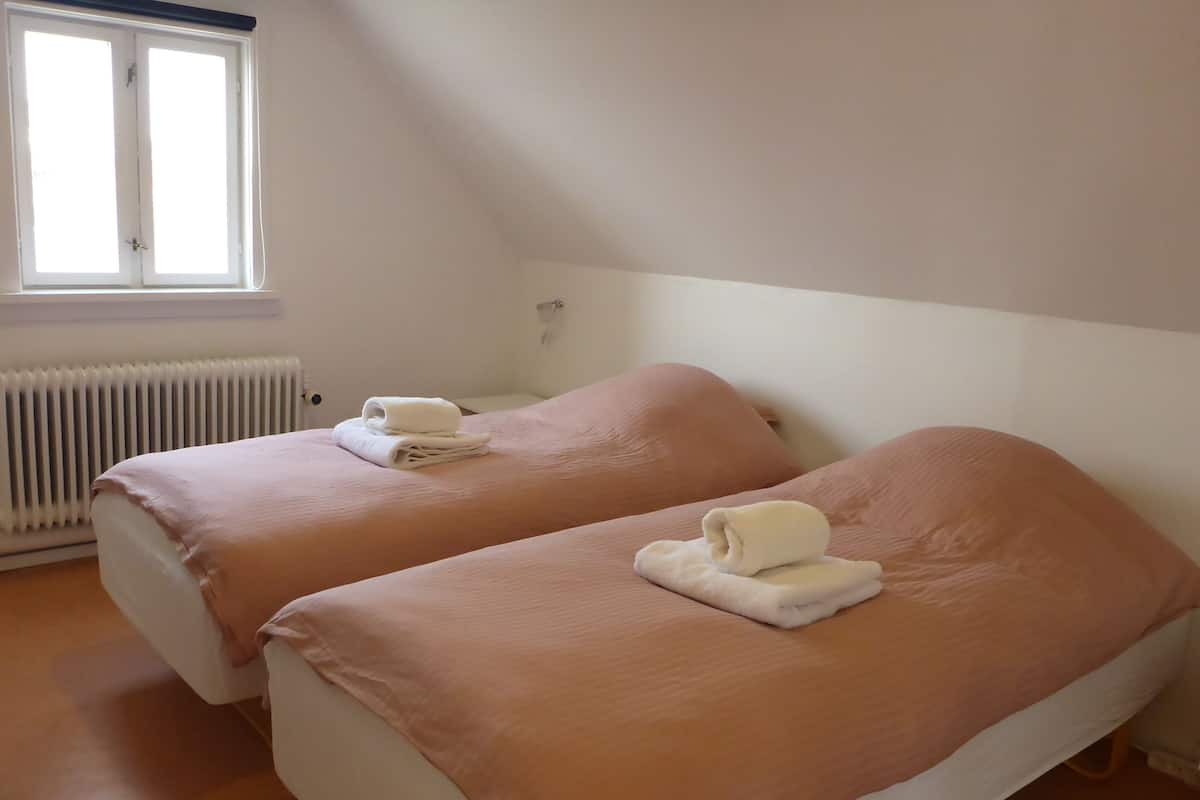 Une chambre avec deux lits, chacun recouvert d’une couvre-lit rose et surmonté d’une serviette blanche pliée. Il y a une fenêtre avec des stores et un radiateur en dessous.