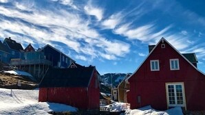 A red building with a gabled roof and a door, surrounded by snow and other buildings. - Café de Upernavik (Upernavik)