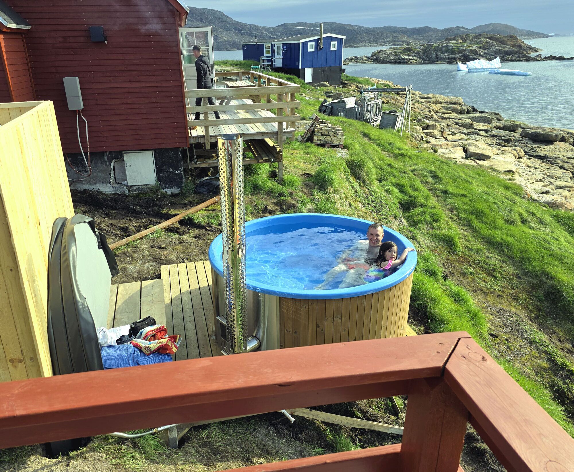 A wooden deck with a hot tub, a red building, and a view of the water and hills.