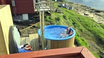A wooden deck with a hot tub, a red building, and a view of the water and hills.