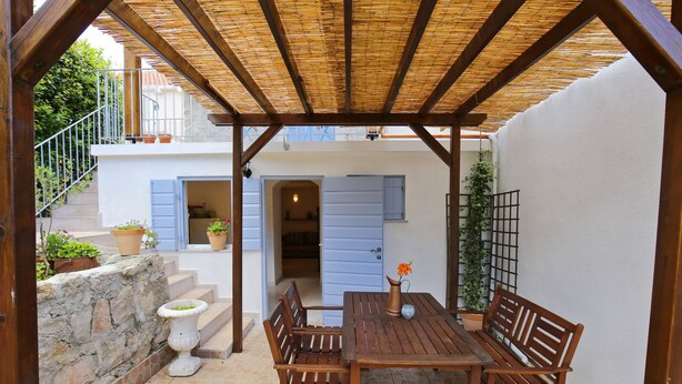 A covered patio with a wooden table and chairs, stone steps, and potted plants. - Stone House Apartment (Preko)
