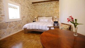 A bedroom with a stone wall, wooden floor, a bed with white bedding, two bedside lamps, and a wooden table with a vase of flowers. - Stone House Apartment (Preko)