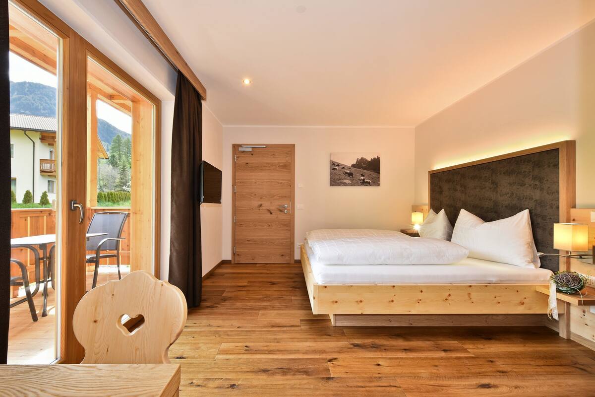 Ein Schlafzimmer mit einem hölzernen Bett, einem Holzstuhl, einer Holztür und Blick ins Freie.