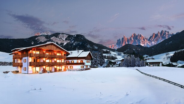 A large, multi-story wooden building with balconies, surrounded by snow-covered landscape and mountains in the background.
