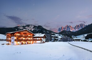 A large, multi-story wooden building with balconies, surrounded by snow-covered landscape and mountains in the background. - Proihof (Funes)