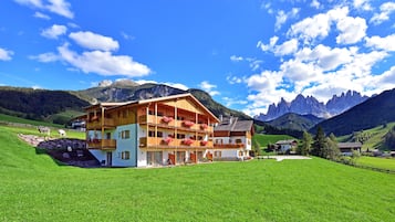A large wooden chalet with multiple balconies, set in a lush green valley with mountains in the background.