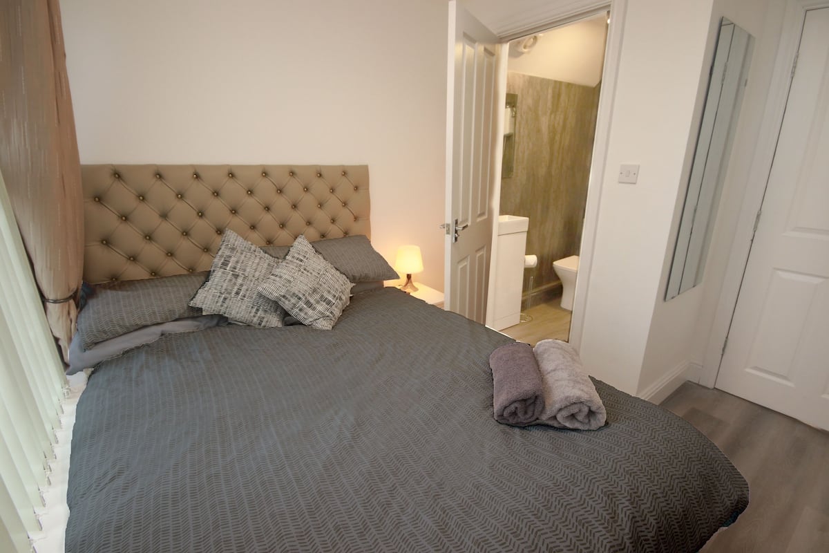 Une chambre à coucher avec une tête de lit capitonnée, un lit avec une literie grise, deux oreillers et une serviette enroulée posée sur le lit.