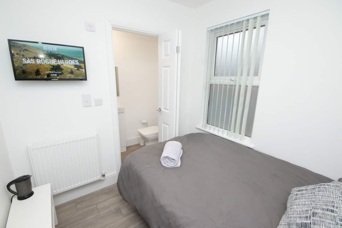 Une chambre avec un lit gris, une serviette enroulée, un téléviseur fixé au mur, un radiateur, une fenêtre avec des stores et une porte blanche donnant sur une salle de bain.