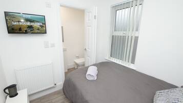 A bedroom with a grey bed, a rolled towel, a TV on the wall, a radiator, a window with blinds, and a white door leading to a bathroom.