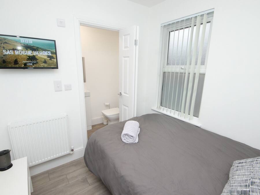 A bedroom with a grey bed, a rolled towel, a TV on the wall, a radiator, a window with blinds, and a white door leading to a bathroom.