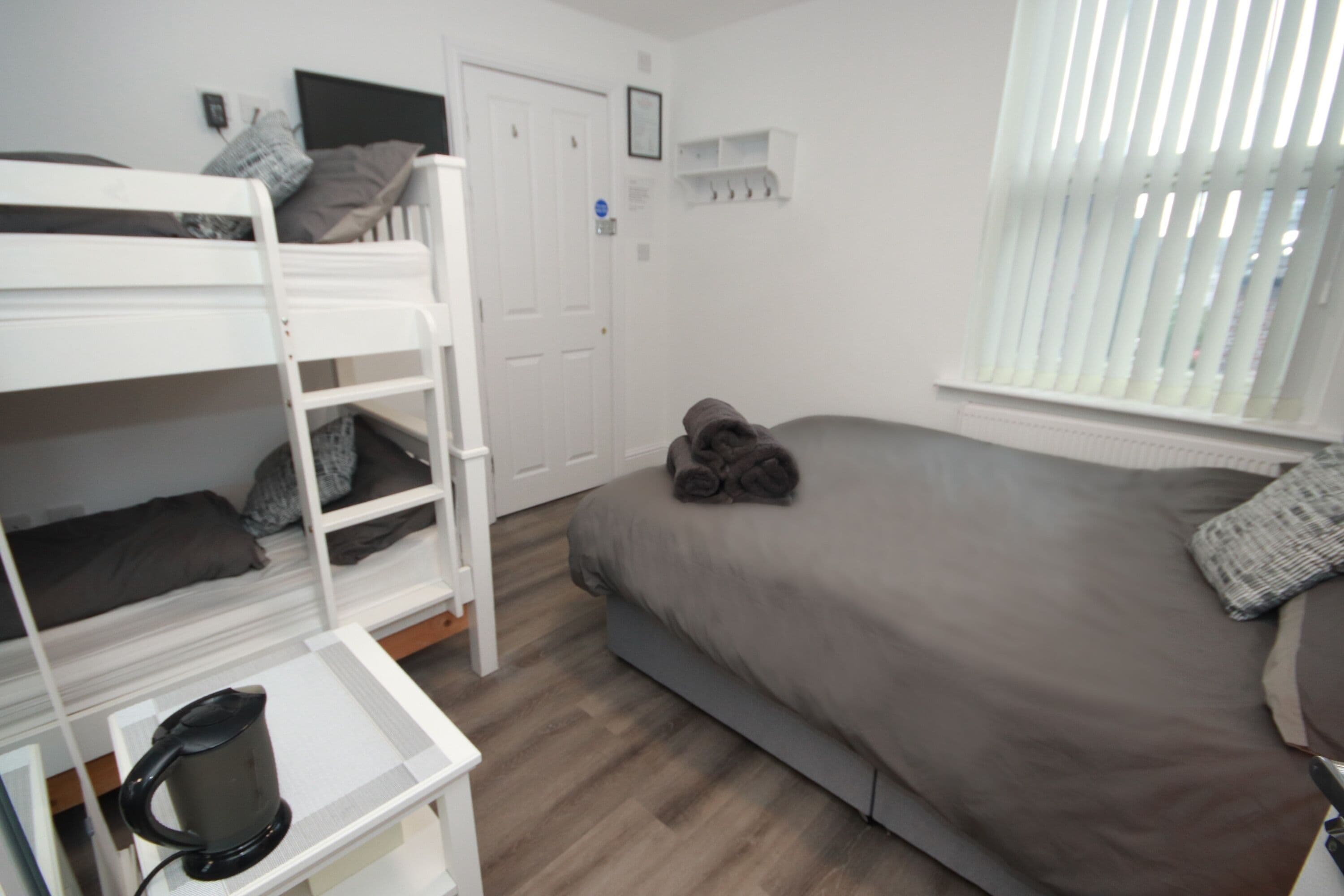 A room with bunk beds, a single bed, a kettle, and a window with blinds.