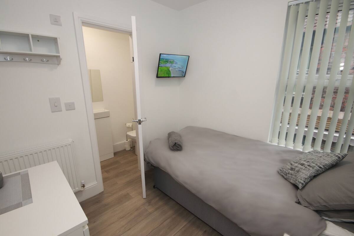 Une chambre à coucher comprenant un lit, une table de chevet, un radiateur, un téléviseur fixé au mur et une fenêtre avec des stores.