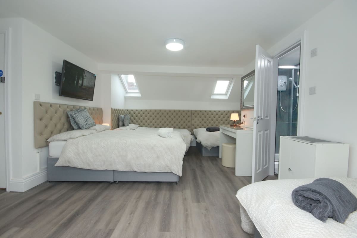 Une chambre avec un grand lit, un bureau et une armoire. Il y a une lucarne au-dessus du lit et un espace douche sur la droite.