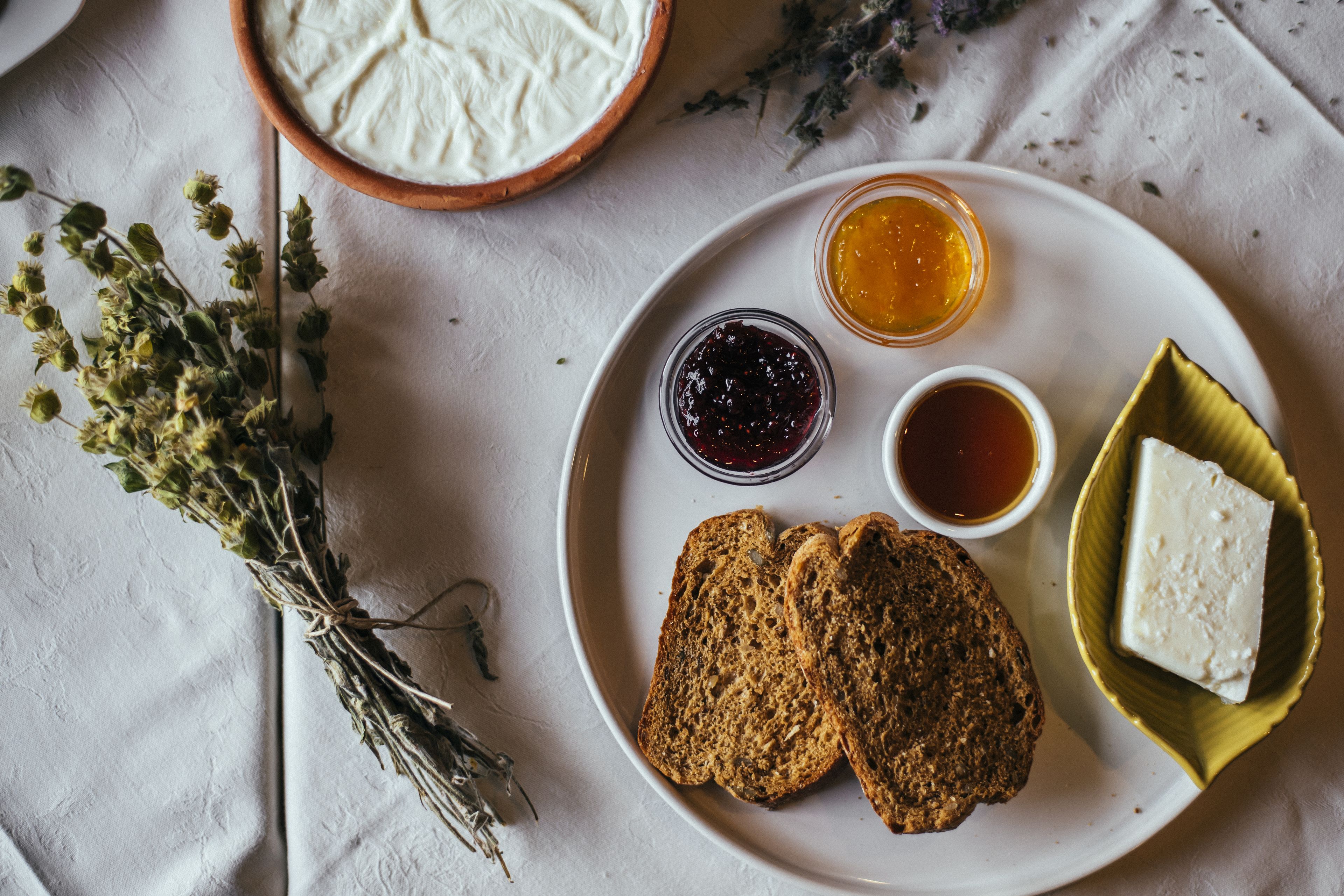 A plate with slices of whole grain bread, a piece of cheese, and two small jars of jam and honey on a white tablecloth.