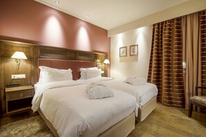Een hotelkamer met twee bedden, een houten hoofdbord, bedlampen, een stoel en een groot raam met gordijnen.