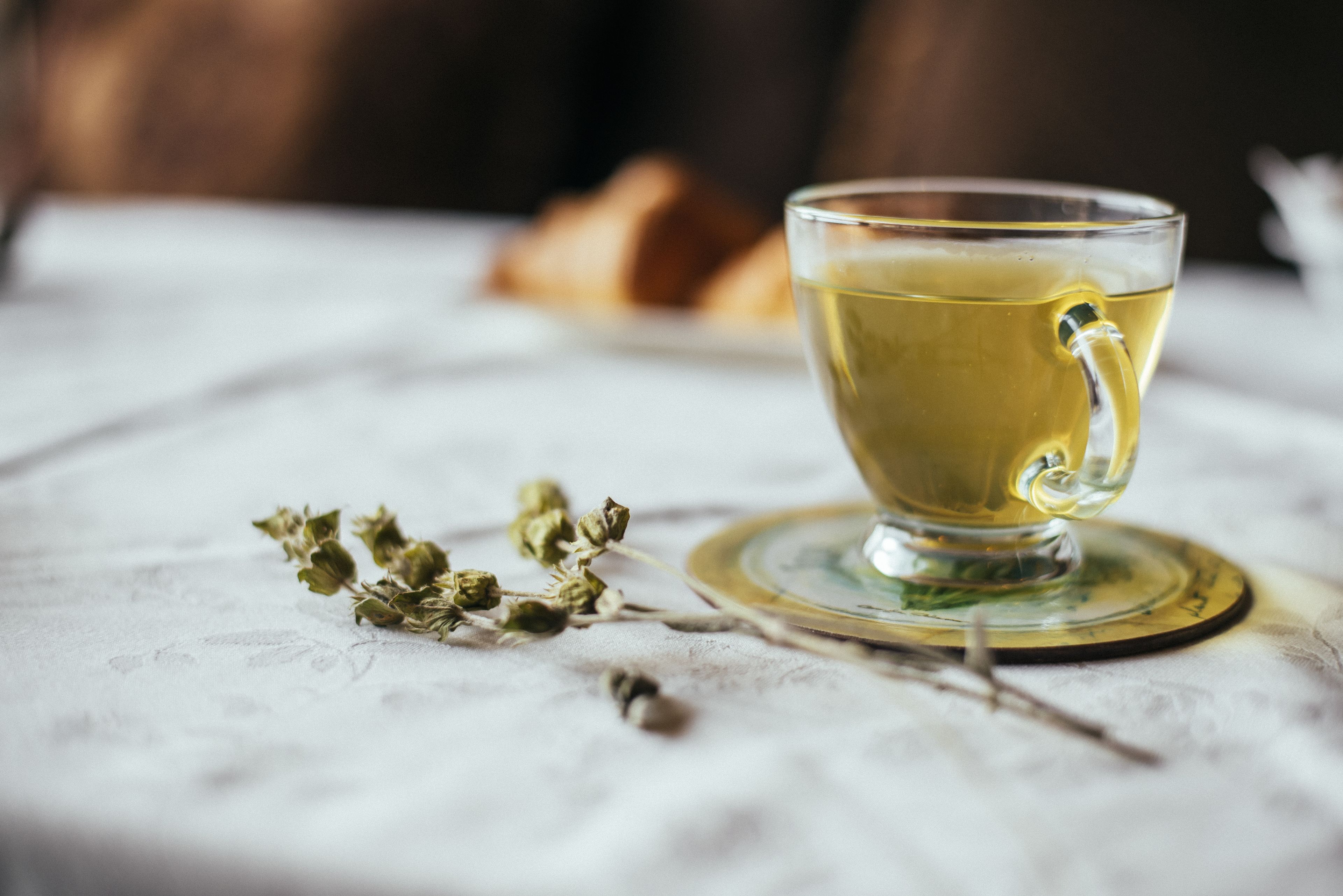 Una taza de té de vidrio transparente sobre un mantel blanco, con un plato debajo, acompañada de hierbas secas.
