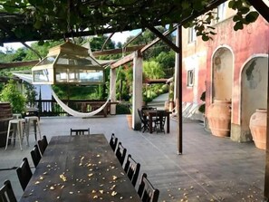 An outdoor dining area with a long table and chairs, a hammock, and a view of a red building with arches. - Agriturismo Gocce di Luna (Celle Ligure)
