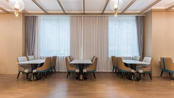 A spacious dining area with a long table and multiple chairs, large windows with sheer curtains, and modern pendant lighting.