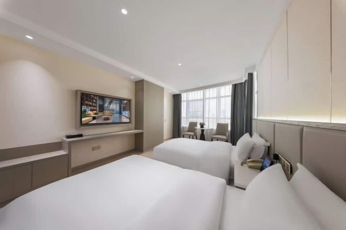 Ein Hotelzimmer mit zwei Betten, einem an der Wand befestigten Fernseher, einem kleinen Tisch und einem Fenster mit Vorhängen.