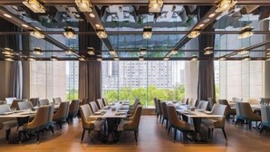 Un espace de restauration moderne, doté de longues tables et de chaises, et de grandes fenêtres offrant une vue sur les bâtiments et la verdure.