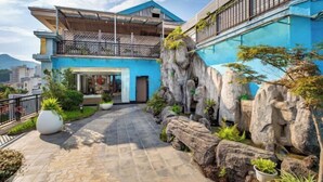 Un bâtiment moderne à deux étages, doté d’une façade bleue, d’un balcon et d’un jardin aménagé avec des rochers et des plantes.