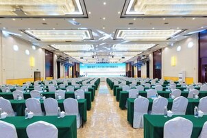 Un salón de conferencias con filas de mesas y sillas, una pantalla grande al frente y un escenario.